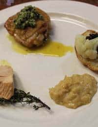 A white plate with a small portion of meat topped with green sauce, a stuffed vegetable with a dark topping, a creamy yellow sauce, and a piece of fish garnished with herbs. The food presentation is arranged around the plate.