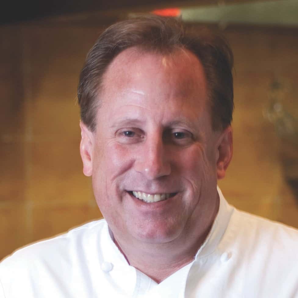 A smiling man with short, light brown hair is wearing a white chef's coat. The background is warmly lit, possibly in a kitchen or restaurant setting.