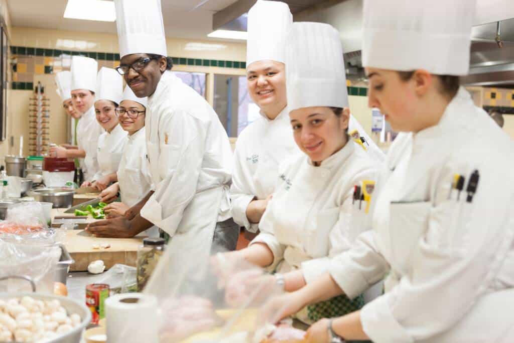 A group of CIA culinary students in white chef uniforms and hats, working together in a kitchen, preparing ingredients.