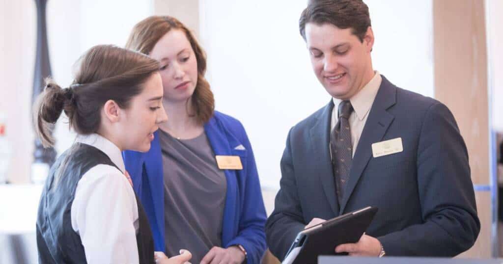 Three professionals are having a discussion. A woman in a vest speaks while another woman in a blue blazer and a man in a suit with a name badge listen attentively, with the man holding a tablet. They appear to be in a workplace setting.