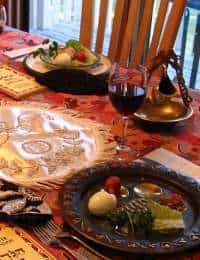 A table is set for a meal, featuring dishes with an arrangement of symbolic foods, a wine glass, and a menorah, suggesting a traditional or holiday setting. The tablecloth is adorned with festive patterns and decorative elements.
