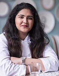 A woman with long dark hair, wearing a white chef's coat, sits at a table with her arms crossed. She has a slight smile on her face. The background features decorative plates on the wall.