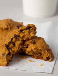A close-up of an orange-tinged cookie with chocolate chips, broken in half, resting on a white napkin with crumbs scattered around. A white cylindrical object, possibly a glass of milk, is blurred in the background.