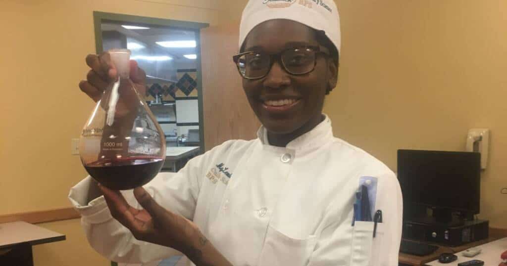 A person wearing a white chef's coat and hat smiles while holding a 1000 ml Erlenmeyer flask containing dark red liquid. They are in a kitchen environment, and there is a computer monitor and phone in the background.