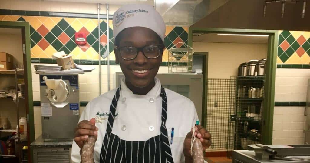 A smiling chef in a kitchen wearing a white chef's hat and coat with a black striped apron holds up two links of sausages. The kitchen has a green and white tiled wall and various kitchen tools and supplies in the background.