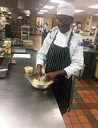 A chef wearing a white hat and apron with black vertical stripes is mixing ingredients in a stainless steel bowl. They are standing in a commercial kitchen, and another chef is visible in the background. Cooking equipment and countertops surround them.