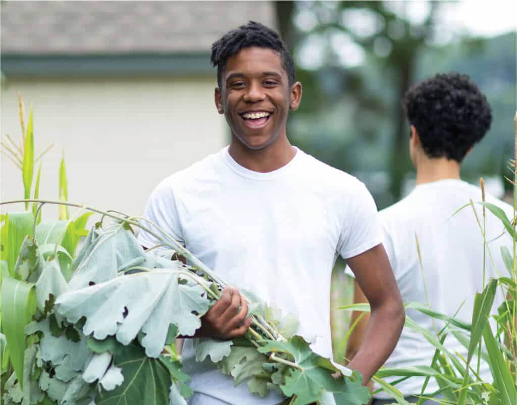 A young man stands outdoors in a garden, smiling and holding large green leaves. He is wearing a white t-shirt. Another person, also in a white t-shirt, is standing nearby with their back turned. The background is blurred, showing greenery and a building.