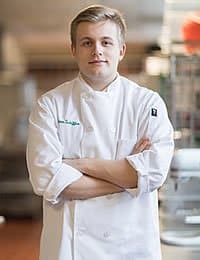 A chef in a white uniform stands with arms crossed in a professional kitchen environment. The background is slightly blurred, showcasing kitchen equipment and a hint of a busy workplace.