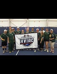 A group of people wearing green shirts and athletic gear stand on an indoor tennis court, proudly holding a banner with the text "HVIAC." The group appears to be a sports team celebrating a victory or achievement.