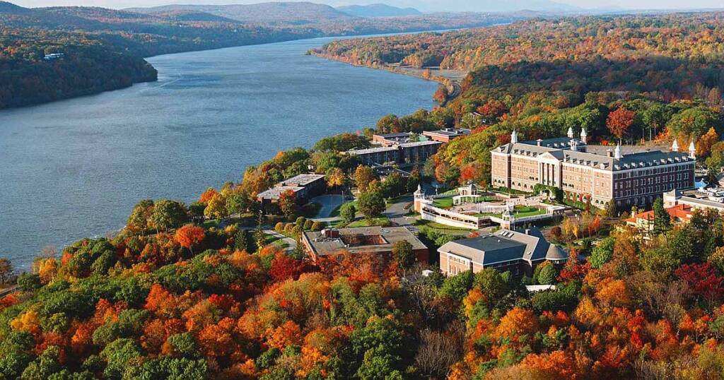 An aerial view of a college campus situated along a river, surrounded by dense forests displaying vibrant autumn colors. The campus buildings are spread out with open green spaces and walking paths. Rolling hills and distant mountains are visible in the background.