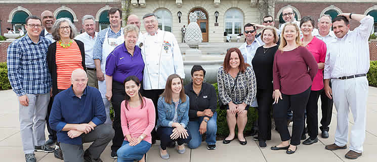 A diverse group of twenty-one adults, men and women, pose together for a group photo outdoors. They are smiling and standing in front of a decorative building with arches and a partially visible garden. One person in the middle is dressed in a white chef’s coat.