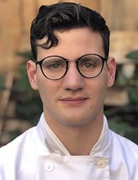 A person with short, dark, wavy hair and glasses is dressed in a white chef's jacket. The individual is looking directly at the camera, and the background appears to be an outdoor setting with plants and wooden elements.