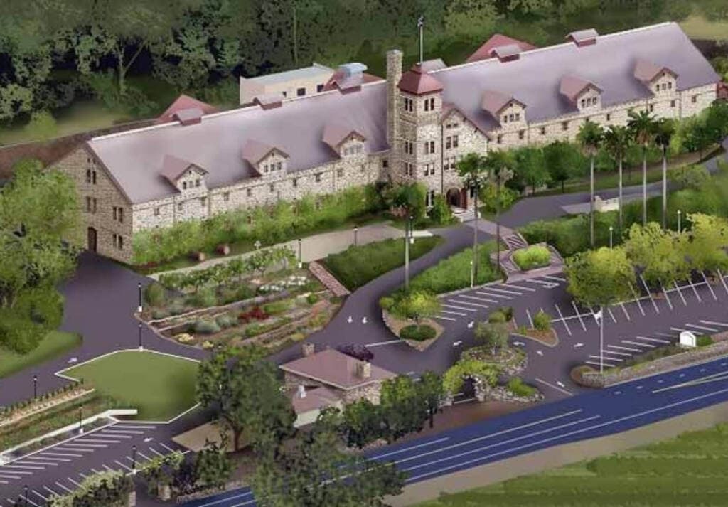 Aerial view of a large, historic stone building with a tower, surrounded by lush greenery and landscaped gardens. The building has a red-tiled roof and several parking spaces. There are palm trees and well-maintained pathways in the vicinity, perfect for CIA Greystone Tours to explore.