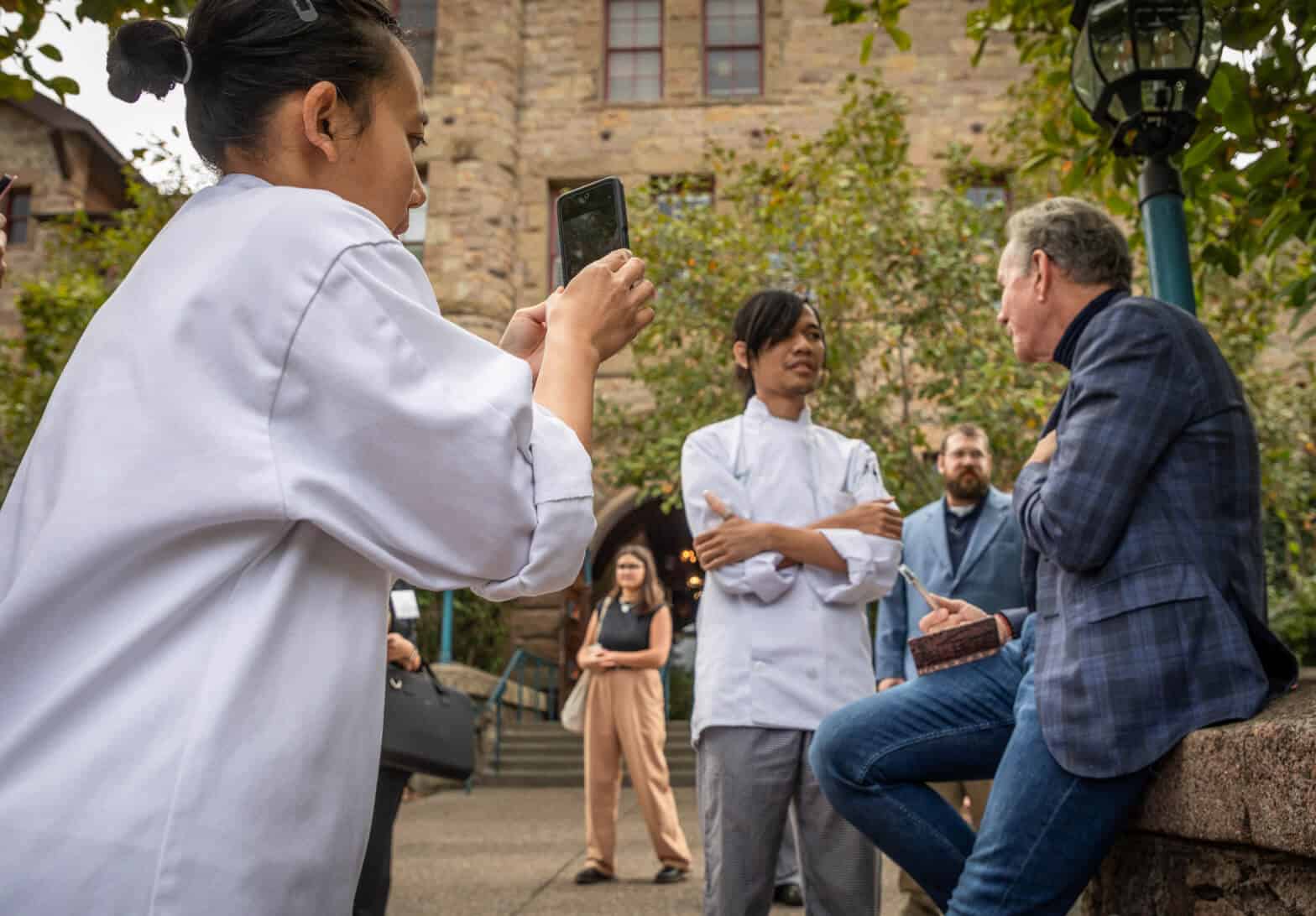 A woman in a white chef jacket takes a photo with her phone of two men, also in chef jackets, conversing with a man in a blue checkered jacket. The interaction appears to take place outdoors, possibly during one of the CIA Greystone Tours, in front of a building with stone walls and green foliage.