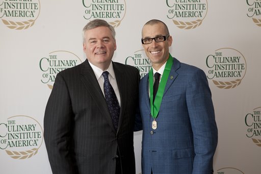 Two men stand in front of a backdrop with "The Culinary Institute of America" logo repeated. The man on the left wears a dark suit and tie, while the man on the right wears glasses, a blue suit, and a medal with a green ribbon around his neck. Both are smiling.