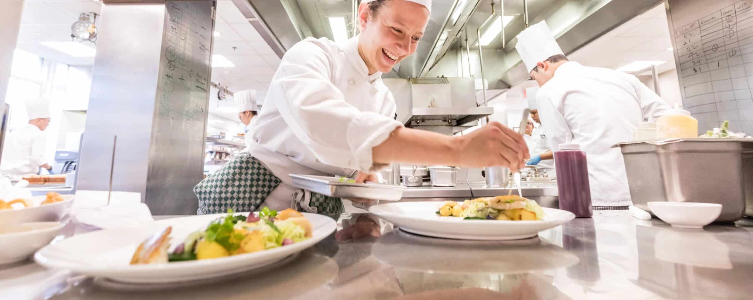 A chef in a professional kitchen garnishes a plated dish while smiling. Other chefs, also in white uniforms and hats, are focused on their tasks in the background. The kitchen workspace is well-lit and equipped with modern stainless steel appliances and counters.