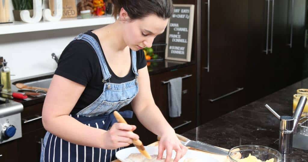 A person with dark hair tied back, wearing a black shirt and striped apron, focuses on brushing a substance onto a food item in a modern kitchen. Shelves with decorative items and kitchen tools are visible in the background.