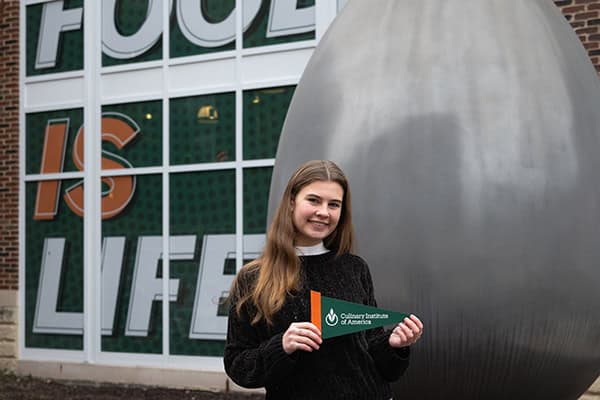 A young woman holding a pennant that reads "Culinary Institute of America" stands smiling in front of a building with green and white signage that reads "IS LIFE." The building, bustling with energy from Accepted Students Day, has a large silver sculpture to the right of the woman.
