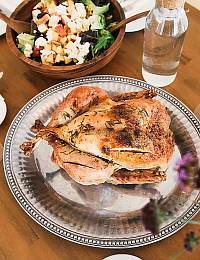 A roasted chicken garnished with herbs on a silver platter is placed on a wooden table. In the background, there is a mixed salad in a wooden bowl and a glass bottle of water. The table is partially decorated with a small vase of flowers.