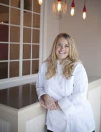 A smiling person with long blonde hair, dressed in a white chef's coat, stands with arms resting on a counter. Behind them, there's a white-paneled wall with a window and hanging light bulbs with red accents. The setting appears to be a cozy, well-lit indoor space.