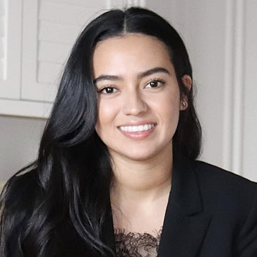 A young woman with long, black hair is smiling at the camera. She is wearing a black blazer with a lace top underneath. The background shows light-colored shutters and a neutral-toned wall.