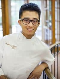 A person with short dark hair and glasses is smiling, leaning against a railing. They are wearing a white chef's jacket embroidered with text on the left side. The background features large windows, suggesting an indoor setting with natural light.