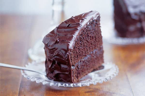 A close-up of a slice of rich, moist chocolate cake with thick, glossy chocolate frosting on a clear glass plate. The chocolate cake has multiple layers with generous amounts of frosting between them. A fork is positioned next to the slice.