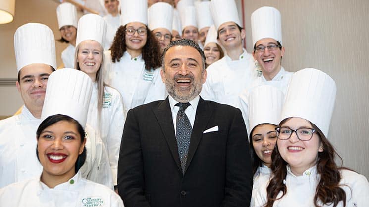 A group of culinary students in white chef uniforms and tall hats stand on stairs, smiling. A man in a suit stands in the center, also smiling. The group is indoors, and the background has plain walls. The mood is cheerful and celebratory.