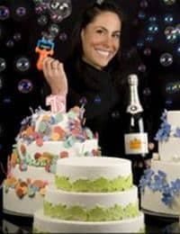 A woman smiles while holding a toy bubble gun surrounded by three decorated cakes and a bottle of champagne. The cakes are adorned with colorful decorations and bubbles fill the background.