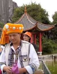 A person in a traditional outfit stands in the foreground with one hand holding a pink object. In the background, there is another person in an orange hat and a red-roofed pagoda surrounded by lush greenery.