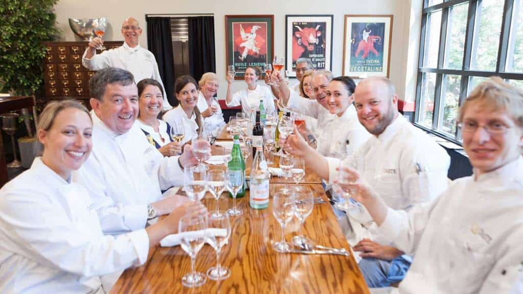 A group of chefs dressed in white coats sit around a wooden table, holding glasses and smiling. The table is set with empty wine glasses, bottles of water, and wine. In the background, there are colorful posters on the wall and large windows letting in natural light.