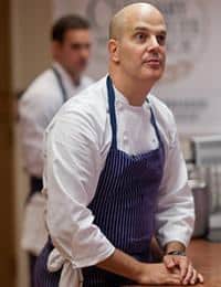 A chef wearing a white coat and a blue striped apron stands at a kitchen counter, looking attentively forward. Another person in a white chef's coat is in the background, slightly out of focus. Both are in a professional kitchen setting.