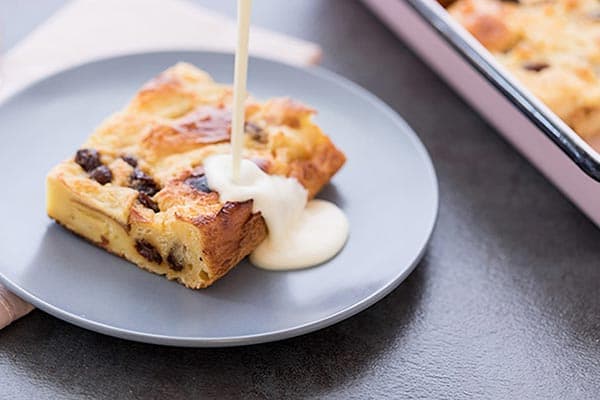 A slice of bread pudding with raisins on a gray plate being topped with a stream of white sauce, likely custard or cream. The plate is set on a dark surface, and part of the baking dish with more bread pudding is visible in the background.