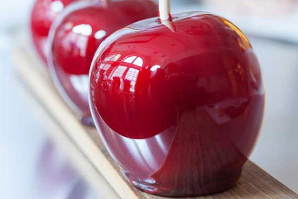 Three shiny, red candy apples are lined up on a wooden plank. The glossy coating reflects light, showcasing their smooth surfaces. The apples are mounted on wooden sticks and have a rich, deep red color.