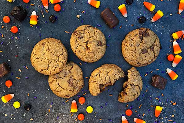 A set of six chocolate chip cookies, one partially eaten, arranged on a dark surface. Surrounding the cookies are colorful candies, including candy corn, chocolate pieces, and round sweets in orange, yellow, and black, as well as scattered purple and orange sprinkles.