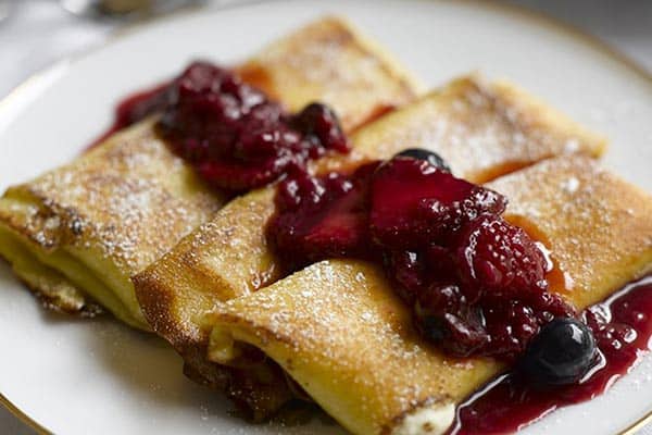 A plate with three rolled crepes topped with powdered sugar and a fruit compote of berries. The compote appears glossy and vibrant, with visible whole berries, adding a rich, colorful contrast to the golden-brown crepes.