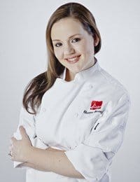 A smiling woman with long brown hair is wearing a white chef's coat with red embroidered lettering and a logo on the left chest. She stands with her arms crossed against a plain white background.