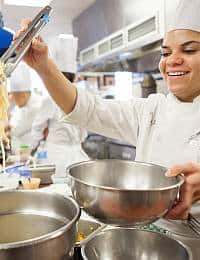 A smiling chef in a white uniform lifts pasta with tongs above a stainless steel bowl in a bustling kitchen with other chefs working in the background.