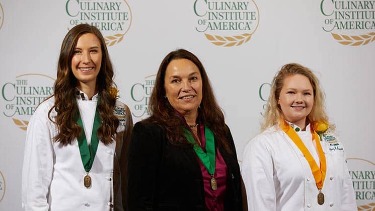 Three women stand against a backdrop featuring "The Culinary Institute of America" logo. The two women on the sides are wearing white chef jackets with medals around their necks, while the woman in the center is in business attire with a green medal and a black blazer.