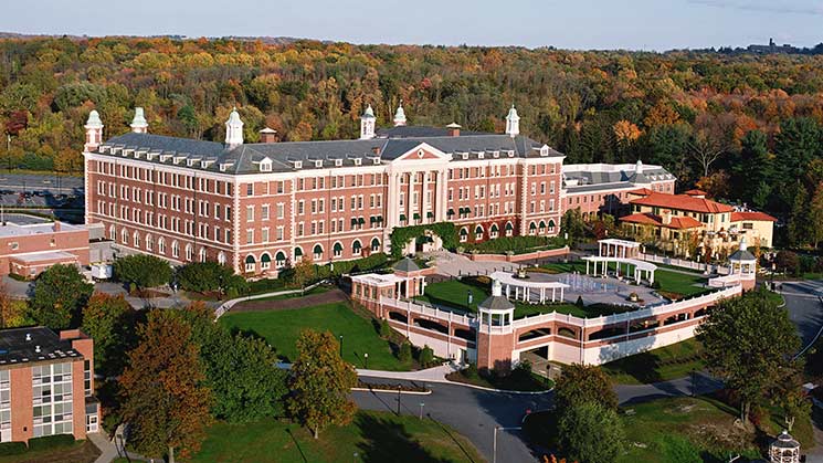 Aerial view of a large, red-brick building with cream trim and white columns, surrounded by lush greenery and colorful autumn trees. The building appears to be part of a larger campus with additional structures and pathways visible.