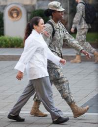 A woman in a white chef's coat and gray pants walks beside a woman in military uniform. They are outdoors, on a paved area with another person in military attire visible in the background.