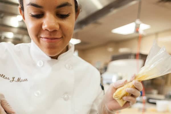 Close-up of a smiling chef in a bright kitchen, wearing a white coat and gloves, holding a piping bag filled with cream or frosting. The chef appears focused on their work, showcasing a sense of pride and dedication.