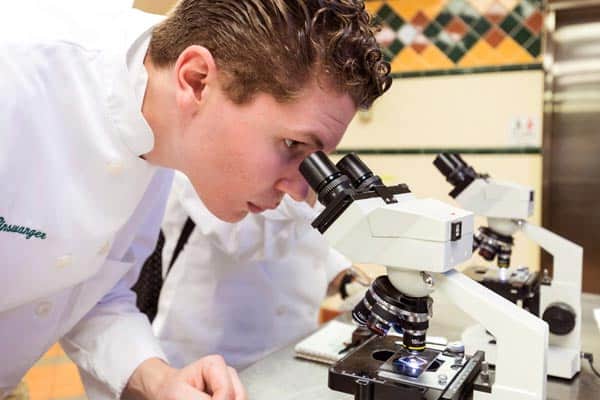 A person wearing a white lab coat closely examines a sample through a microscope in a laboratory setting. Another microscope is visible beside them, and a person in the background can be seen in partial view. The room has tiled walls.