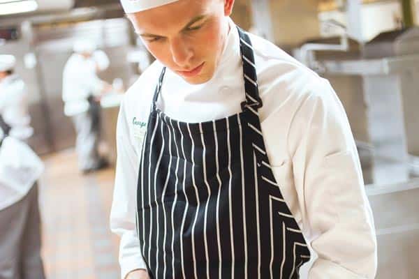 A chef wearing a white uniform, apron, and hat concentrates on preparing food in a professional kitchen. Other chefs are visible in the background, busy with their tasks. The kitchen looks clean and well-equipped.