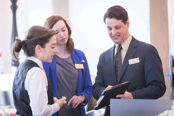 Three professionals are in a discussion. One woman, wearing a white shirt and black vest, is engaged in conversation with a woman in a blue jacket and a man in a suit, who are both looking at a tablet held by the man. They all have name tags.