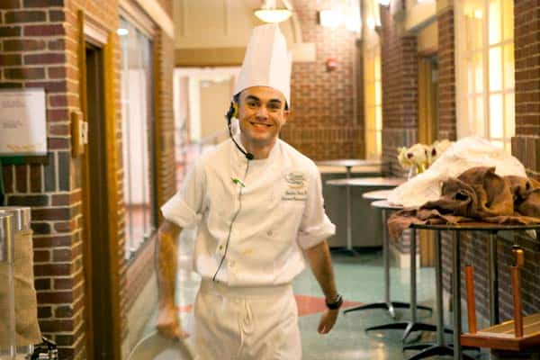 A chef wearing a white uniform and tall white hat smiles and walks down a hallway. He has a headset on and is holding a utensil in his right hand. The hallway has brick walls and is lined with windows. Tables with cloths are visible on the right side.