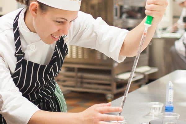 A person in a chef's uniform, including a white hat and striped apron, is smiling and using a pipette to measure and transfer a liquid in a professional kitchen setting. Kitchen equipment and containers are visible in the background.