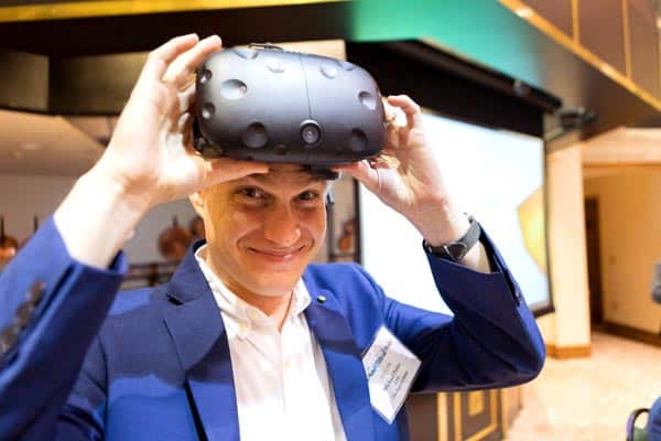 A person in a blue suit and white shirt is smiling while adjusting a virtual reality headset on their head. The background appears to be an indoor setting with a blurred projection screen and other people in the vicinity.