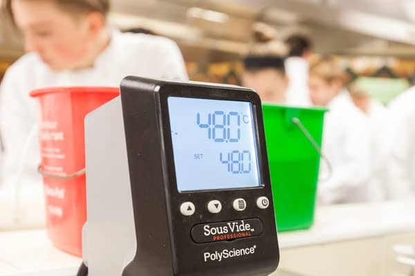 Close-up of a SousVide Professional PolyScience precision cooker showing a temperature reading of 48.0°C with the target set to 48.0°C. Two people in white coats are blurred in the background, along with red and green buckets.