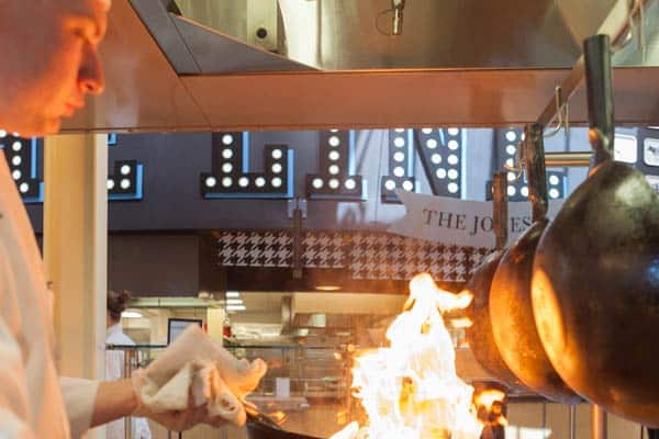A chef in a white uniform is cooking at a stovetop, with visible flames rising from the pan. Above, in the background, there is a sign with the partially visible words "...ING" and another one that appears to say "...THE JOBS...".